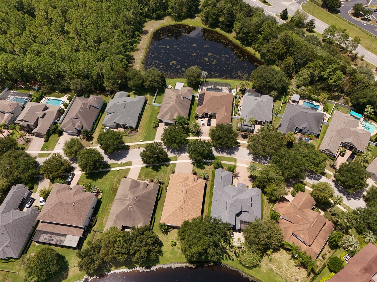 Aerial view of homes along a tree-lined street, with backyard pools and a pond nearby.