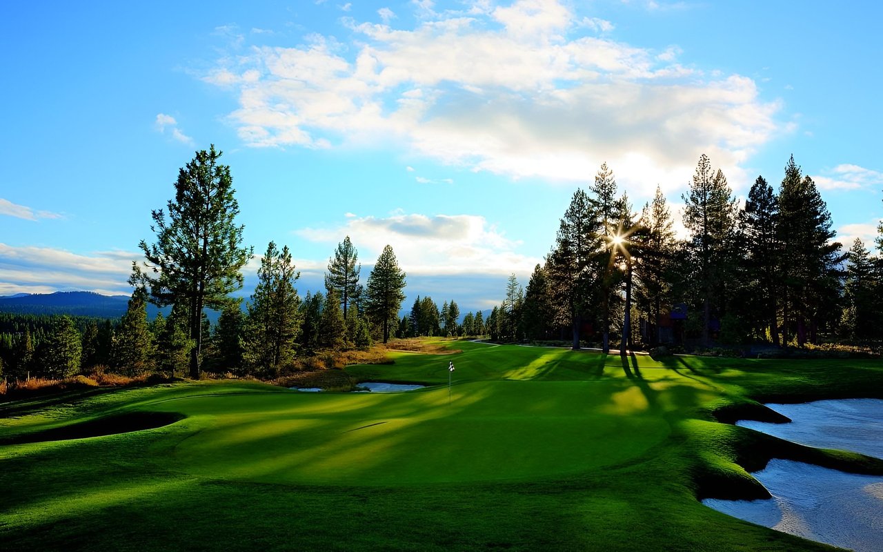 Golf course green with a flagstick, water hazards, and pine trees in golden-hour light.