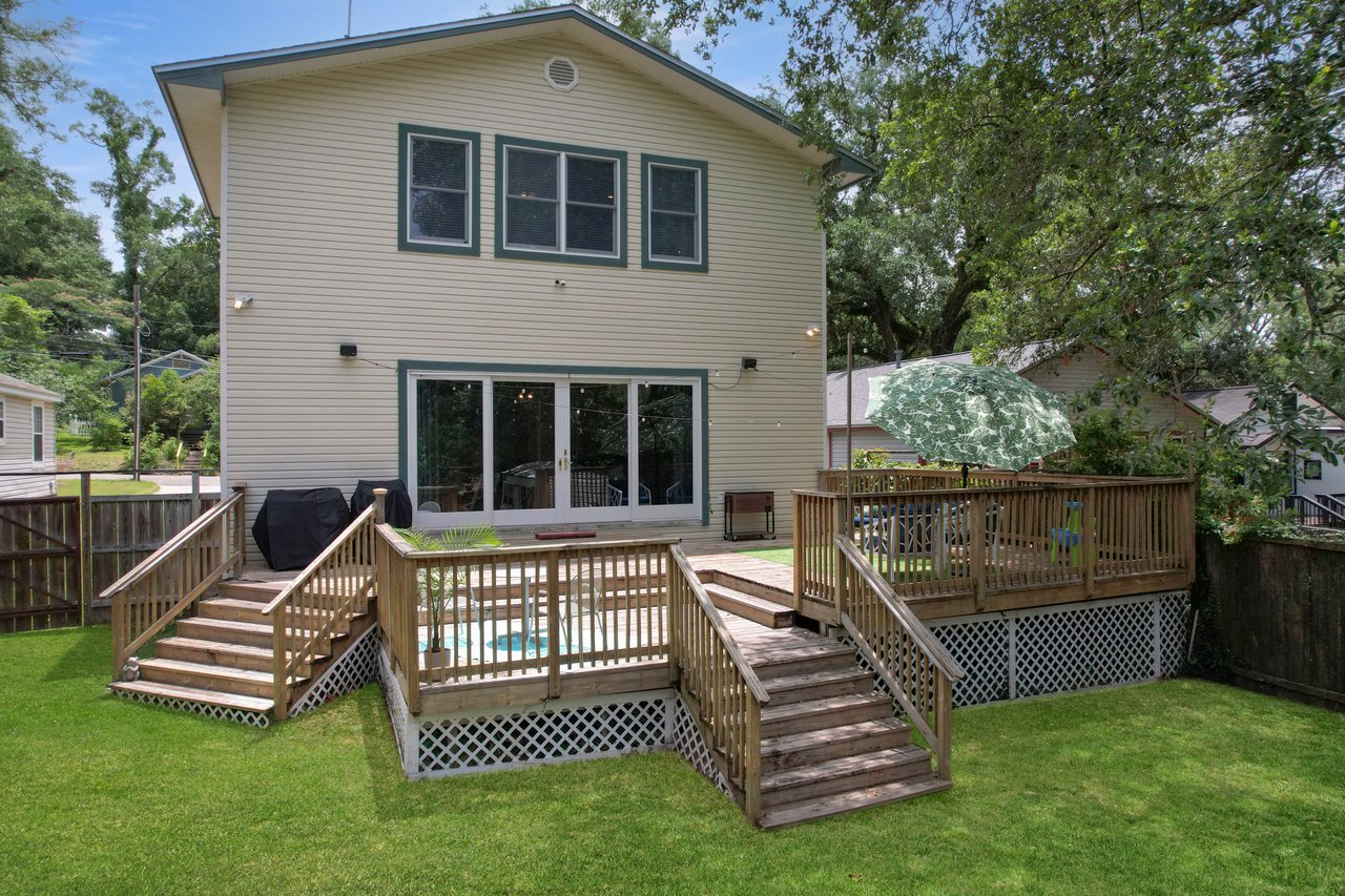 A two-story house with beige siding, large windows, and a wooden deck. Dual staircases lead to a grassy yard, surrounded by trees under a clear sky.