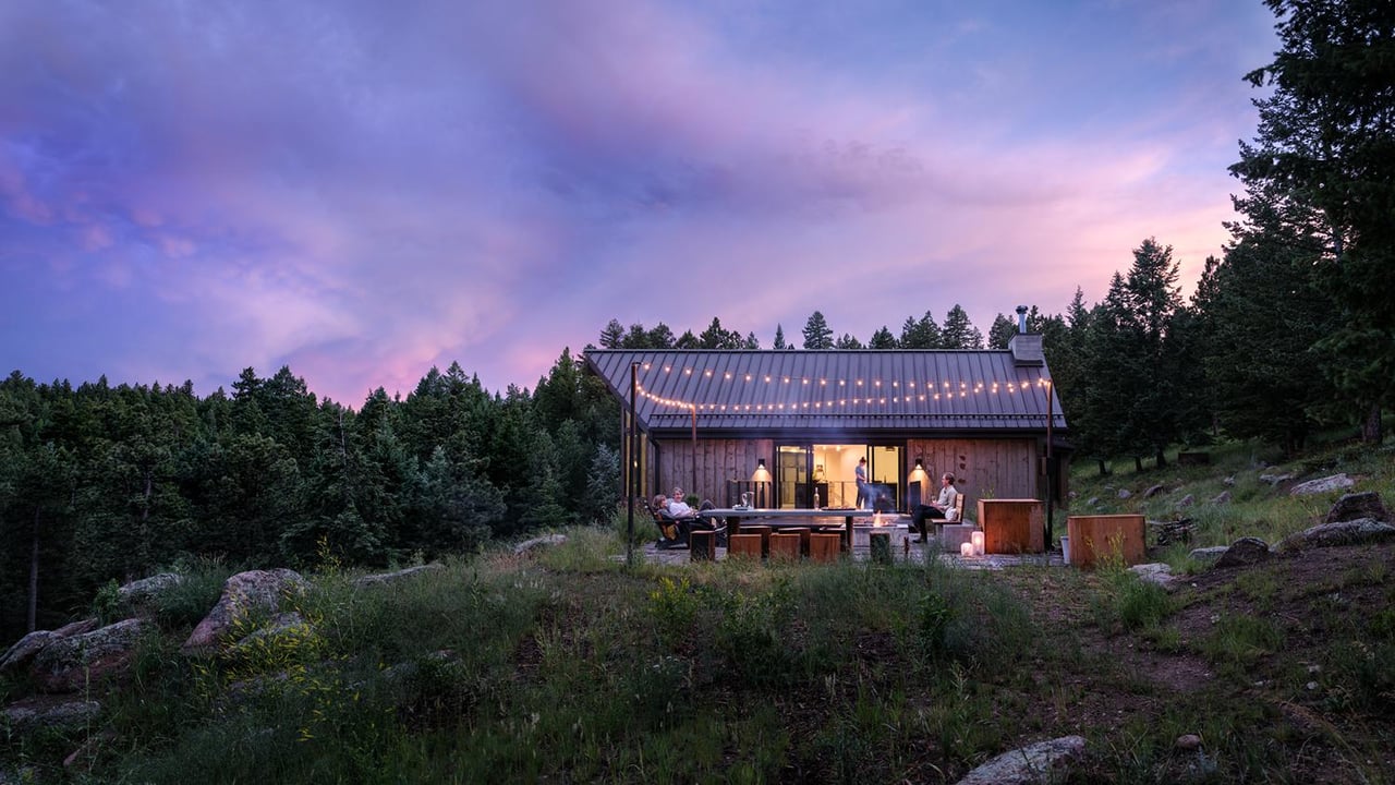 Boulder luxury home's outdoor deck at twilight with string lights, featuring comfortable seating against pine forest backdrop
