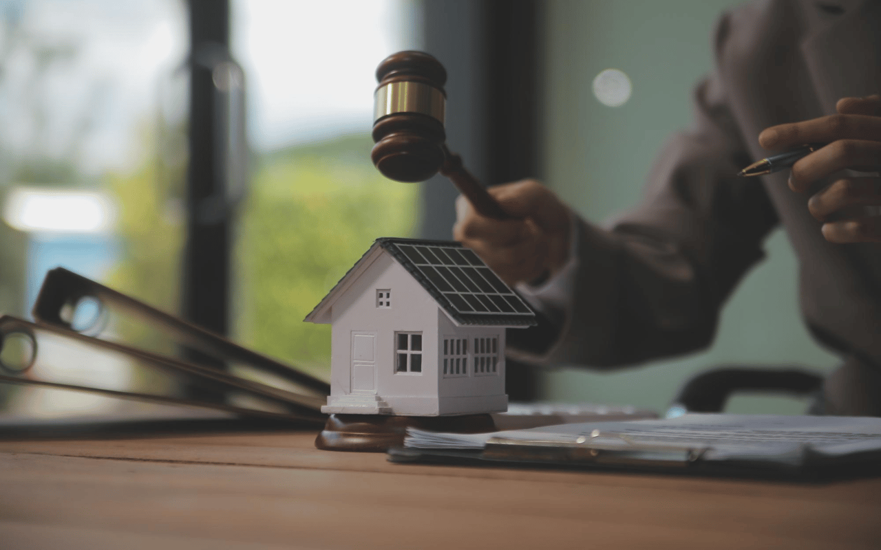 A hand raises a gavel over a miniature solar-panel house and offer papers.