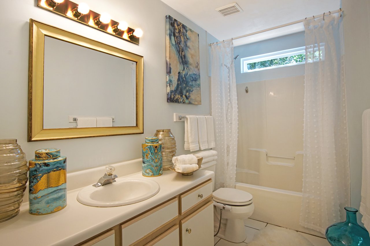Elegant bathroom with a large, gold-framed mirror above a sink, flanked by decorative vases. Soft lighting, white shower curtains, and plush towels create a cozy atmosphere.