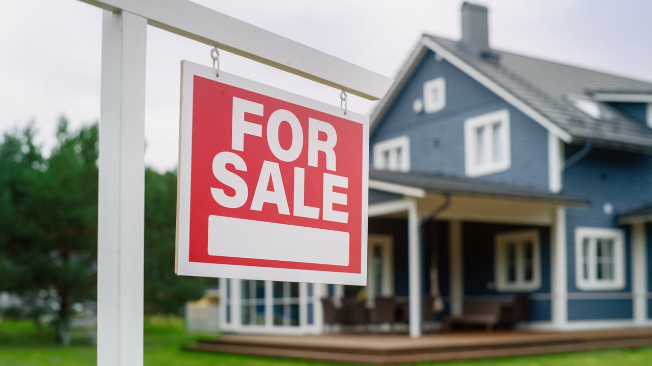 real-estate-pricing-psychology-close-up-of-a-red-and-white-for-sale-sign-in-front-of-a-big-blue-house