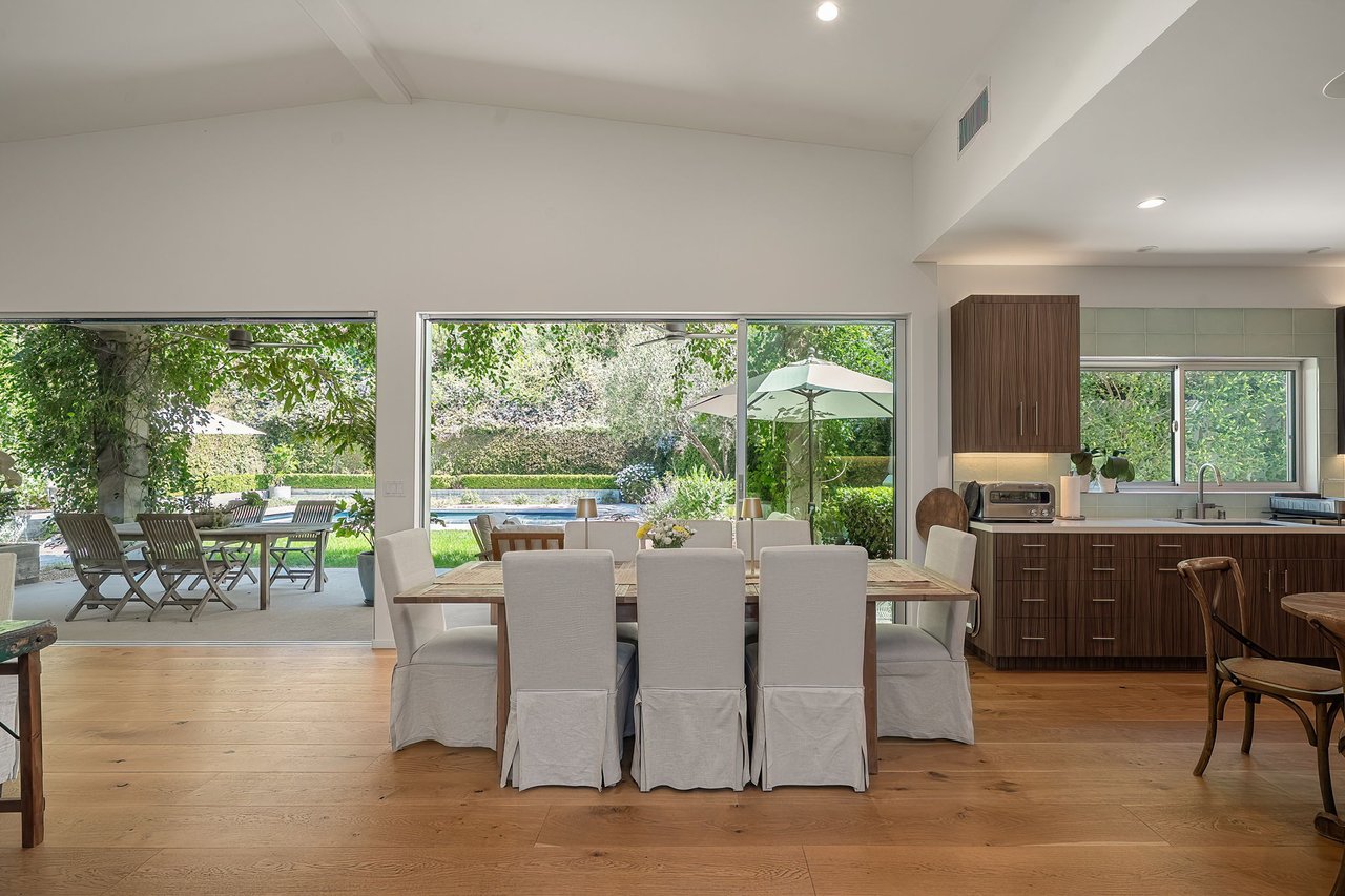 Dining area with white chairs and wide windows overlooking a landscaped poolside yard.