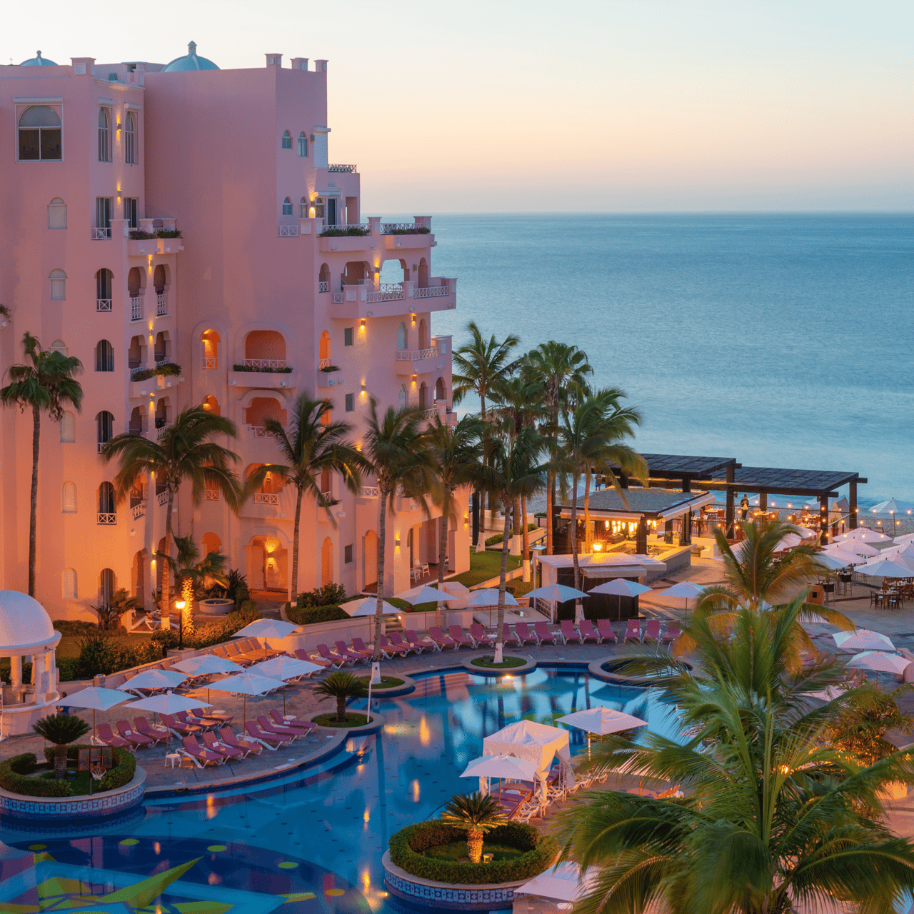 Oceanfront residence with a pink facade, palm-lined pool, and illuminated terrace at dusk.