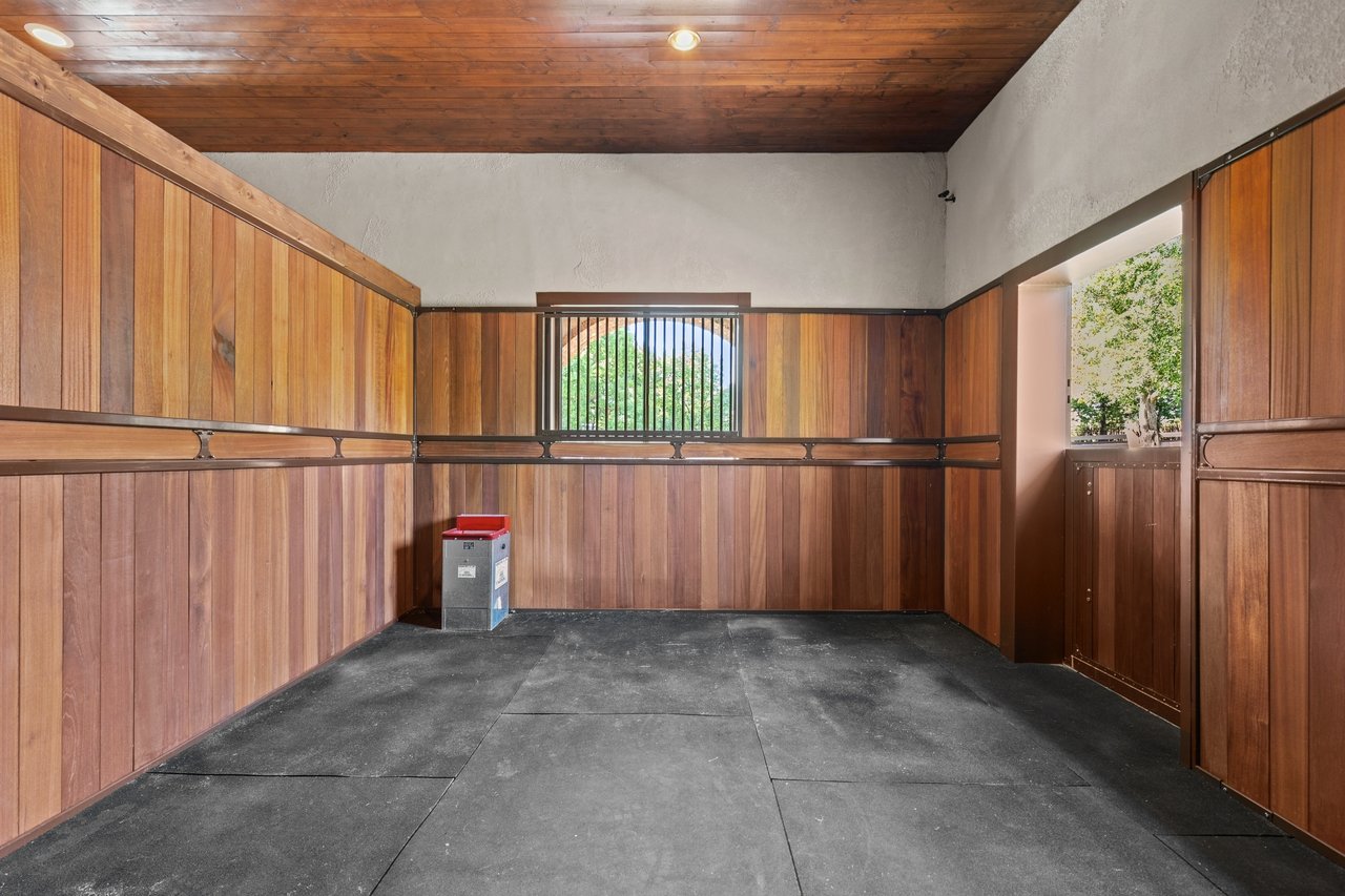 Empty horse stall with wood-paneled walls, rubber flooring, and a barred window.