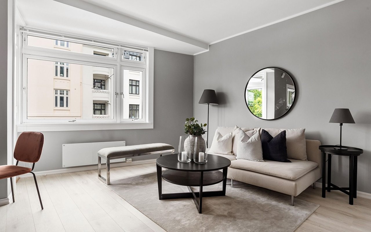 Staged living room with a beige sofa, round coffee table, and large windows overlooking neighboring buildings.