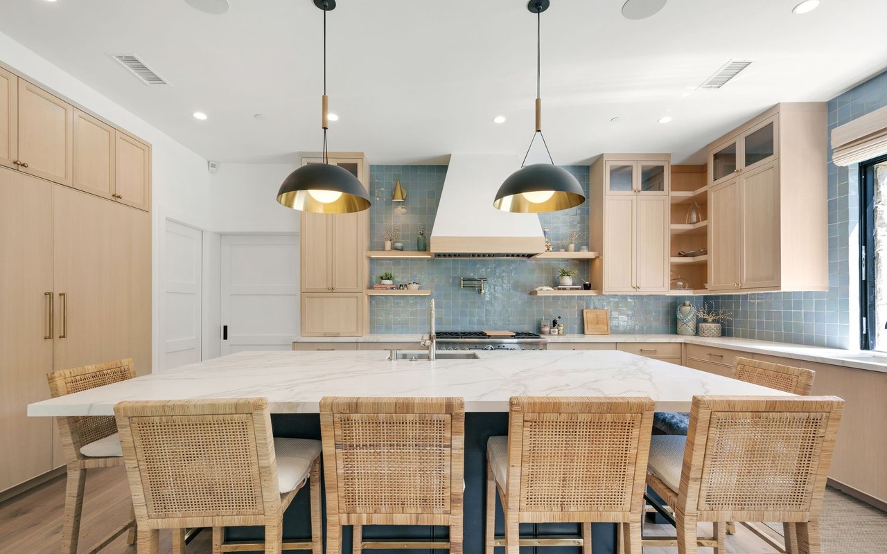Kitchen with a marble island, pale wood cabinetry, blue tile backsplash, and woven bar stools.
