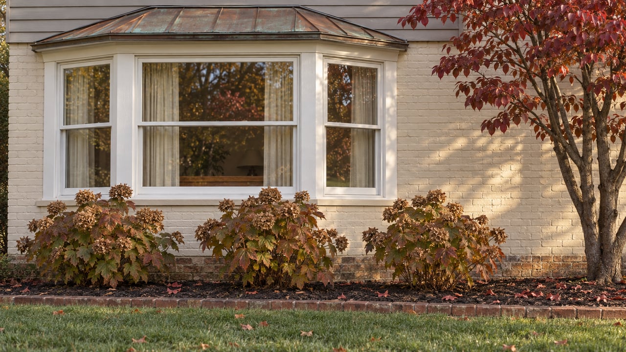 Three-window bay on an established home with painted brick, taupe siding, an aged copper roof and burgundy autumn planting.