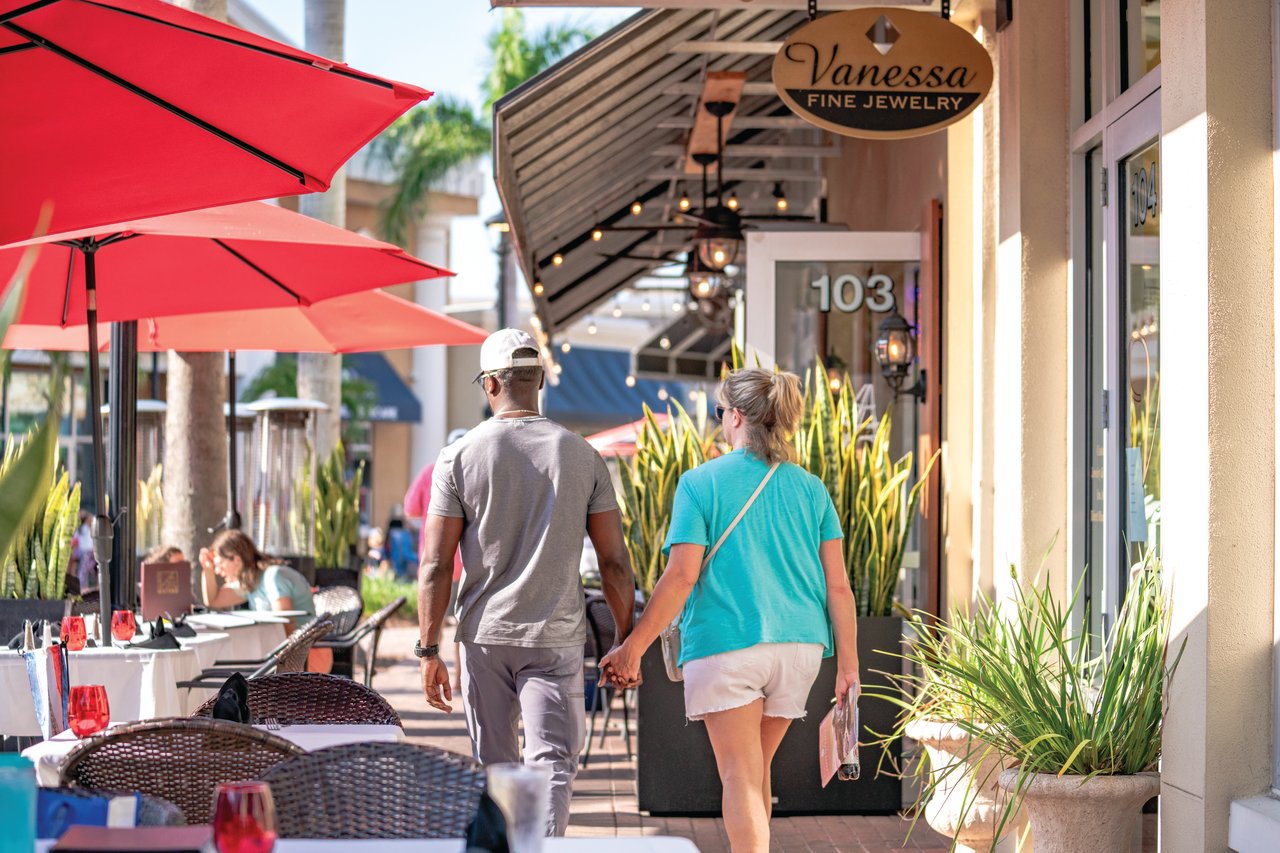 Pedestrians stroll past Vanessa Fine Jewelry and outdoor dining beneath red umbrellas.