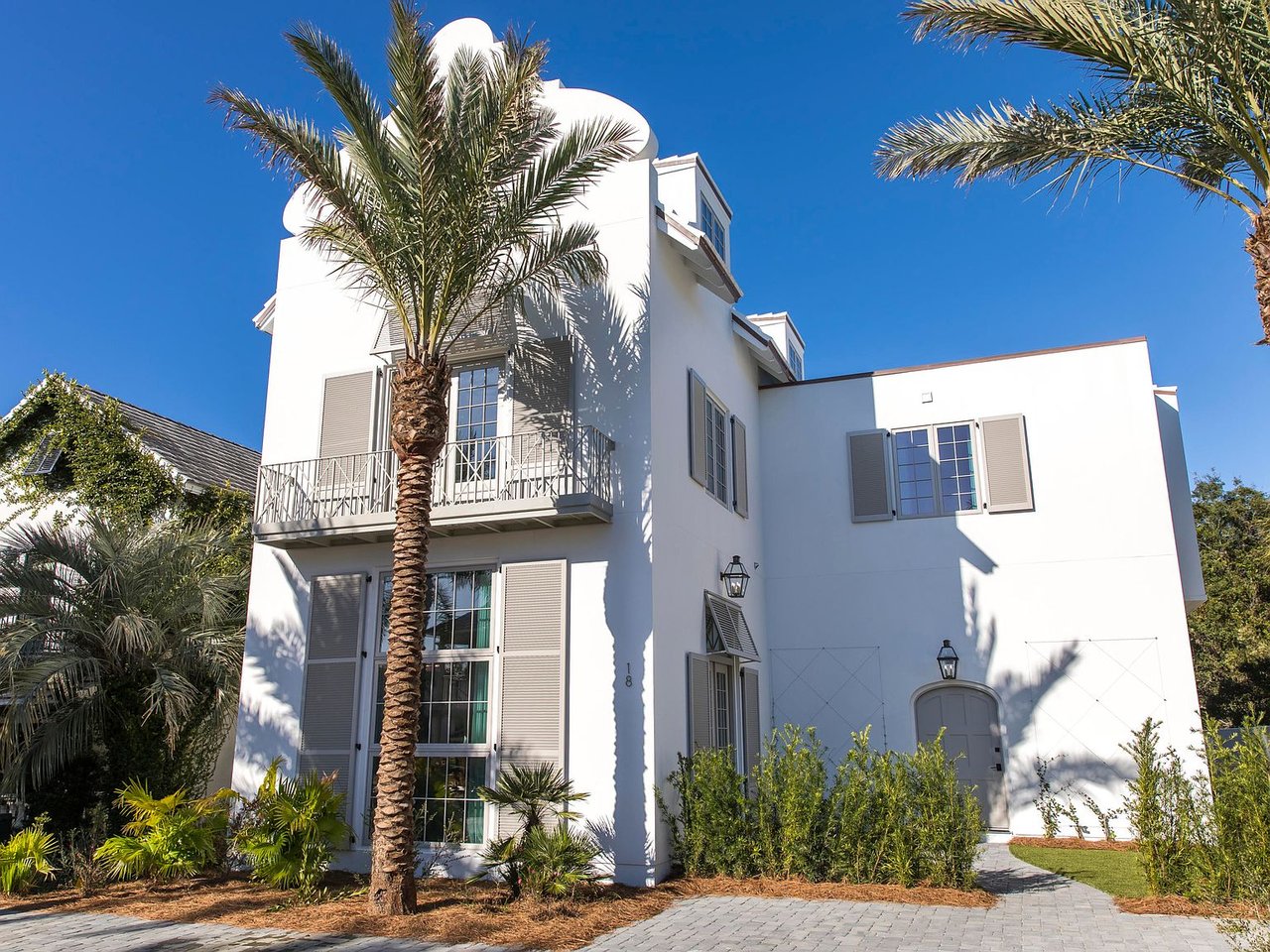 White multi-story home with rounded roofline, balcony, shuttered windows, and palm trees.