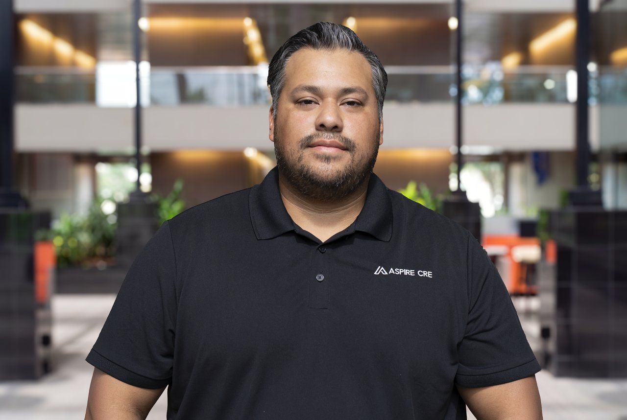 Isaac Puente in a black ASPIRE CRE polo shirt, standing in an office lobby.