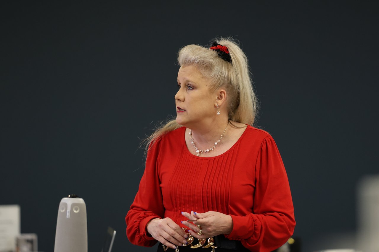 Woman in a red blouse presenting, with blonde hair in a high ponytail and hands together.