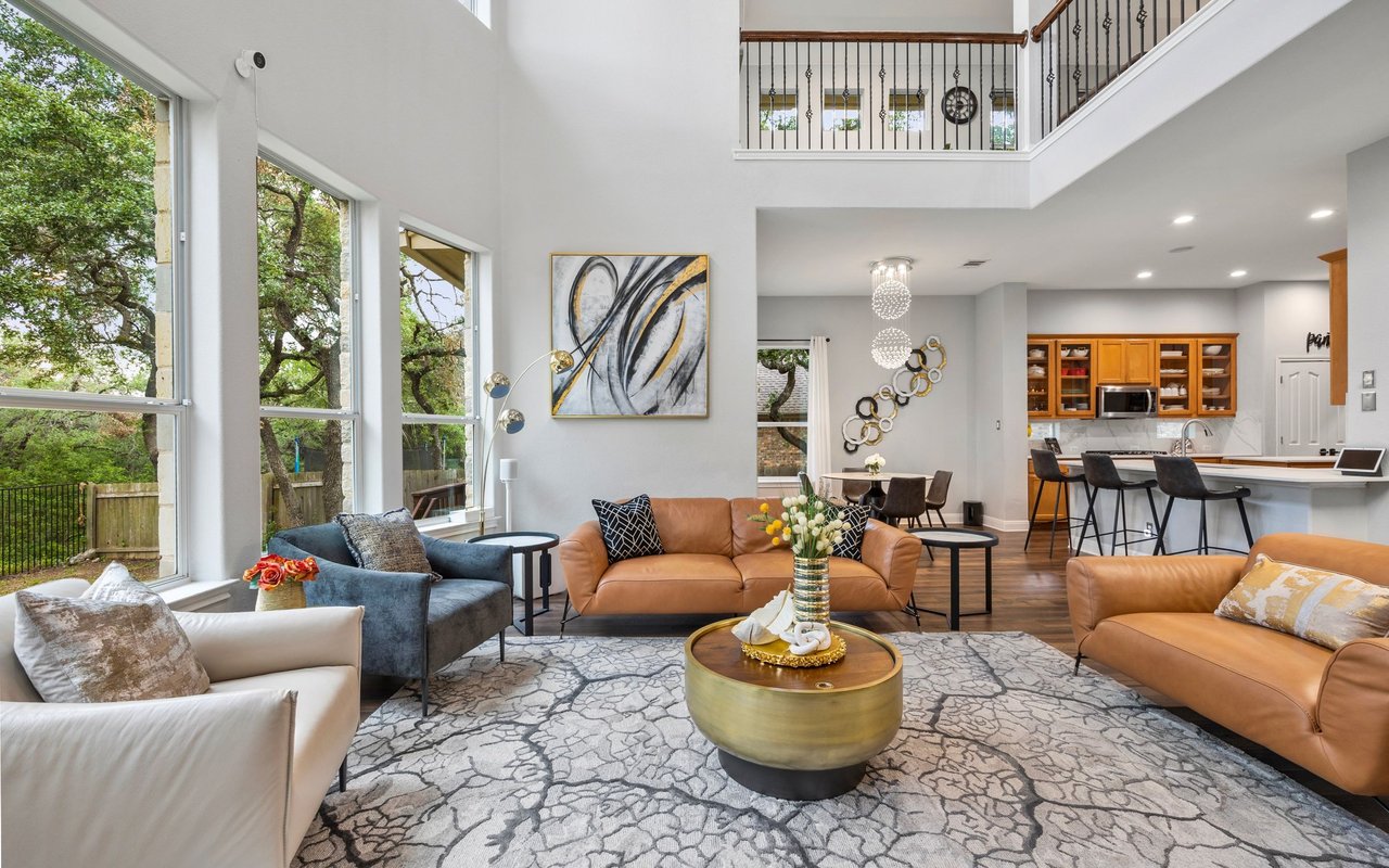 Staged double-height living room with large windows, leather sofas, patterned rug, and open kitchen.