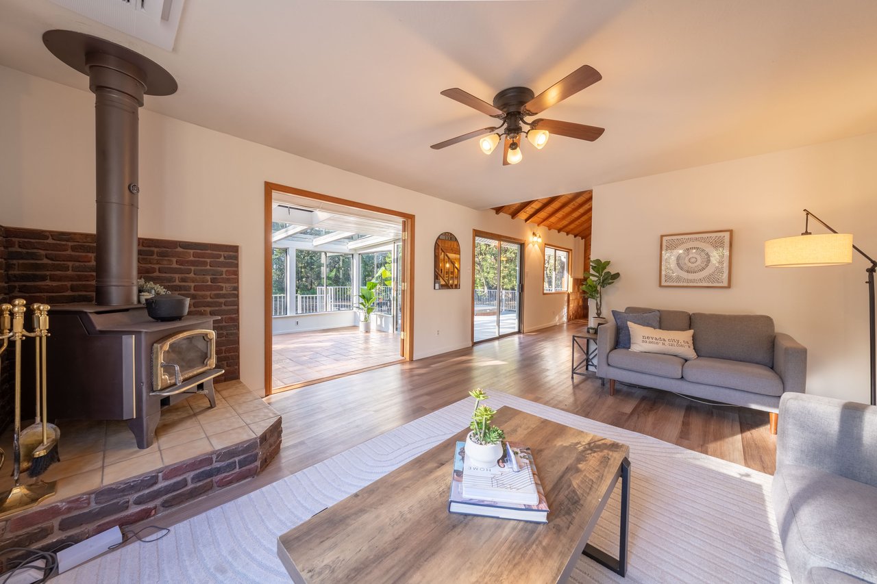 Living room with a brick-backed wood stove, gray sofa, and wide opening to a bright sunroom.