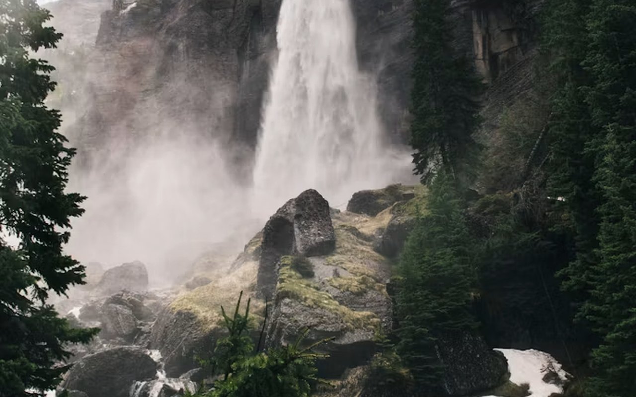 Waterfall cascading over a rocky cliff into a misty basin among evergreen trees.
