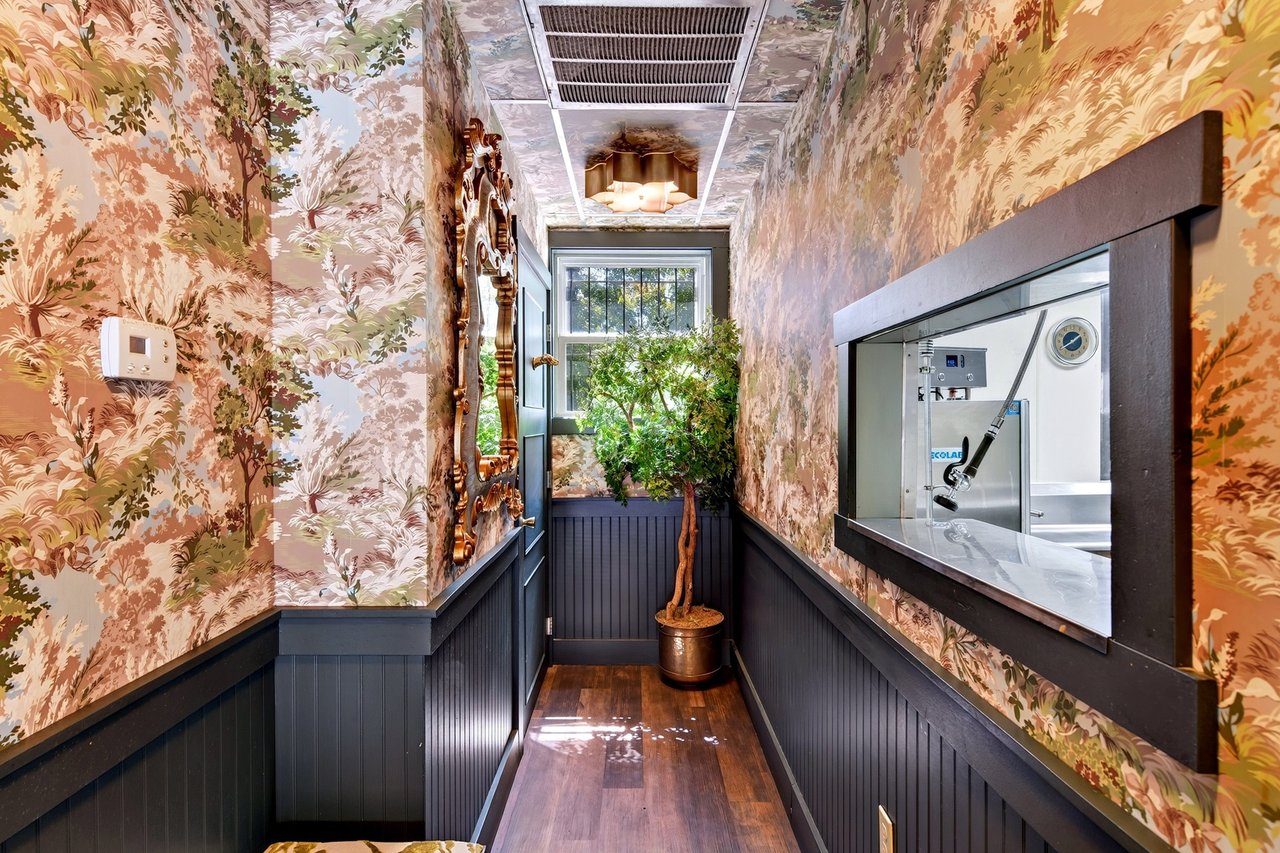 Restaurant hallway with floral wallpaper, dark wainscoting, a kitchen service window, and a potted tree.