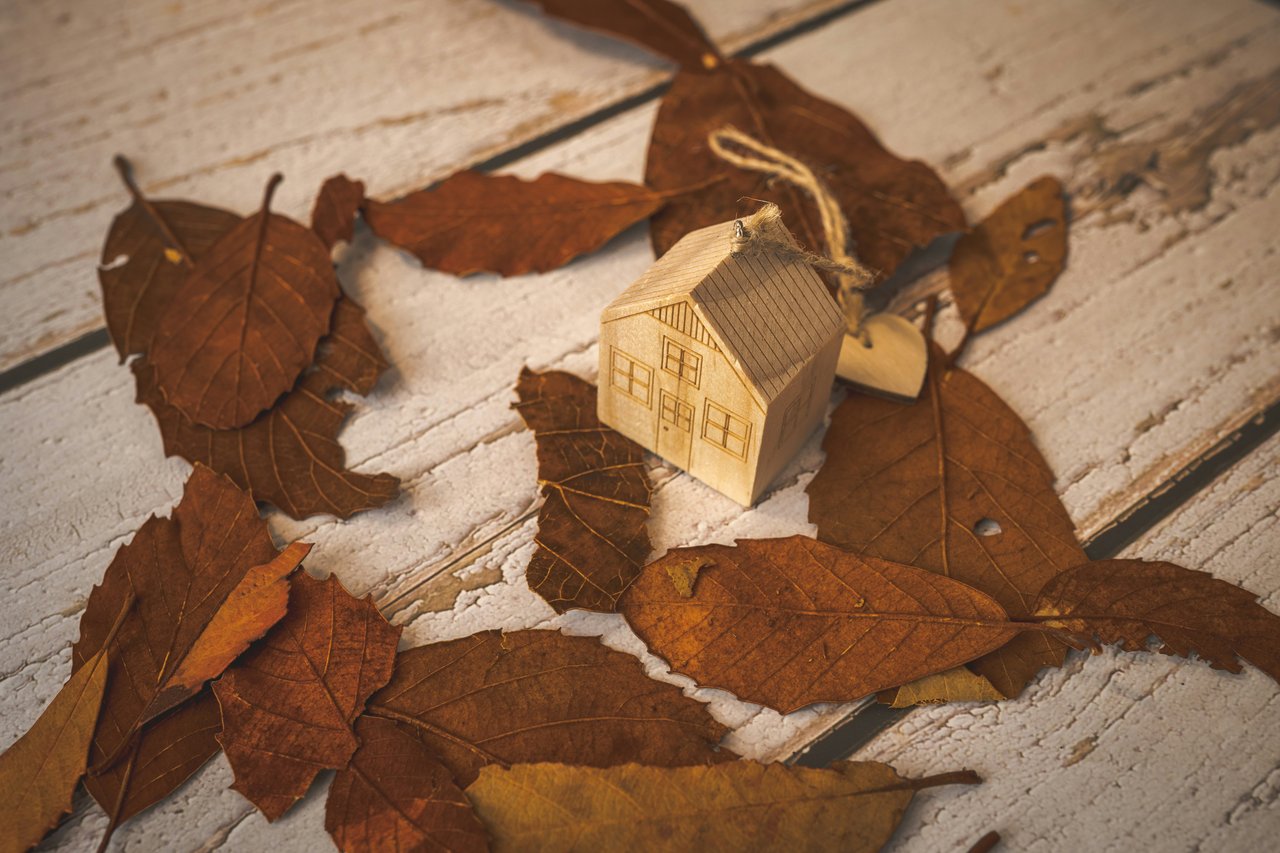 Wooden model house nestled among dry autumn leaves on a white wooden surface.