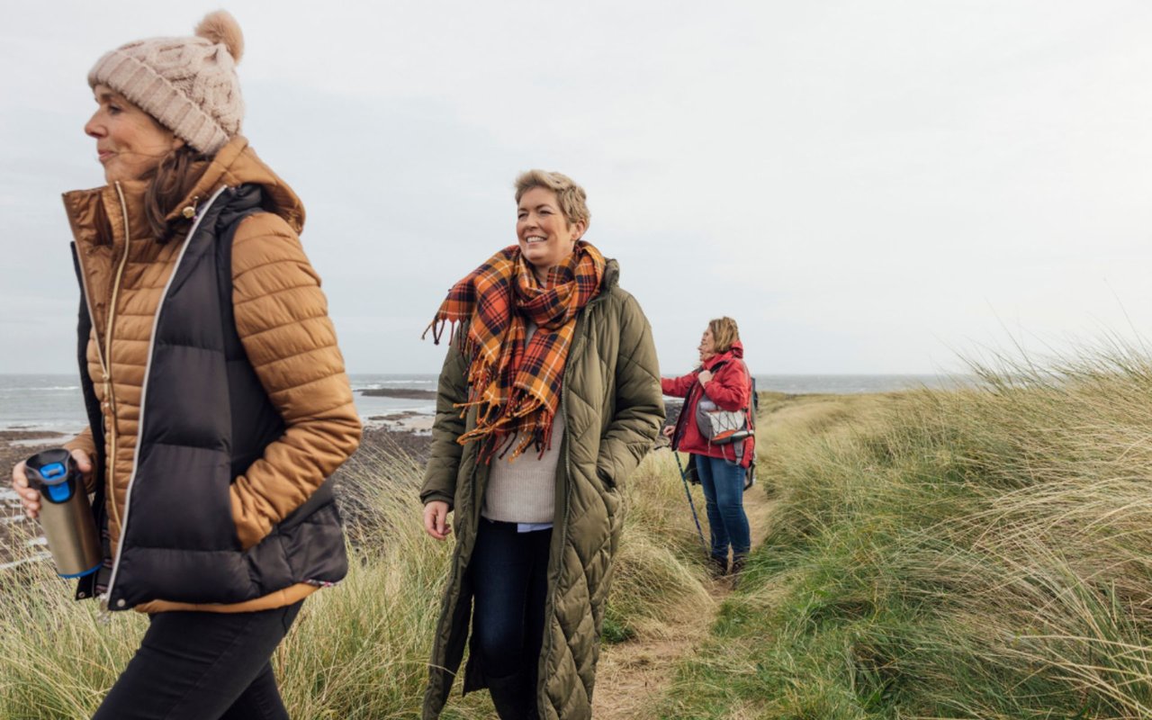 Women are walking through tall grass by the beach.