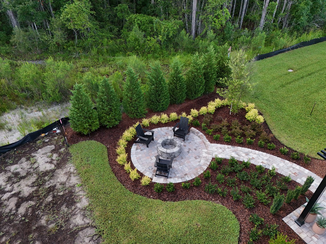 Backyard patio with a circular stone fire pit, Adirondack chairs, and landscaped garden beds.