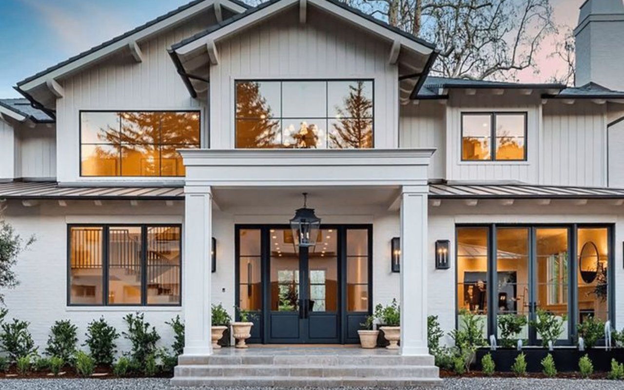 Two-story white house with a covered entry and warm lights glowing through black-framed windows.