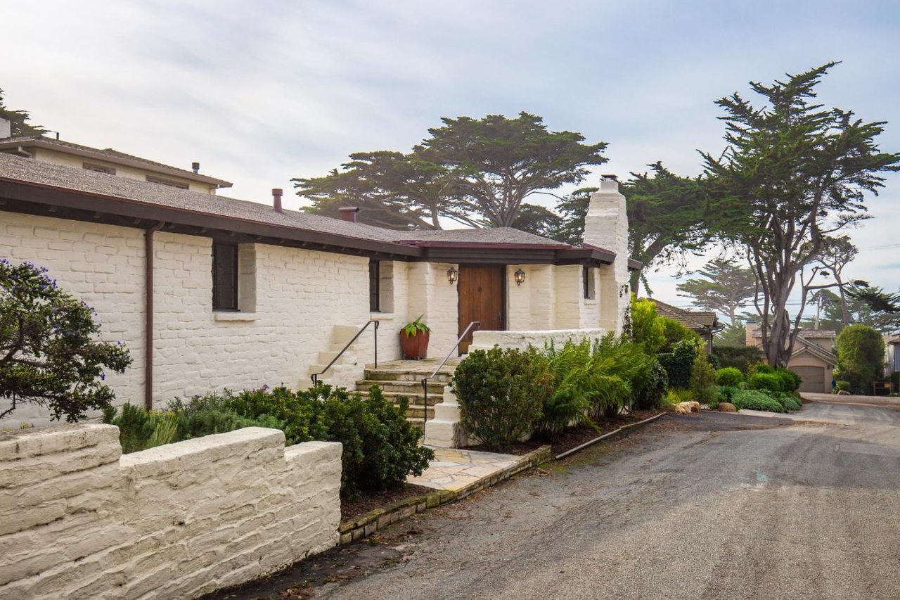 White stone home with a wooden entry, landscaped steps, and tall coastal trees.