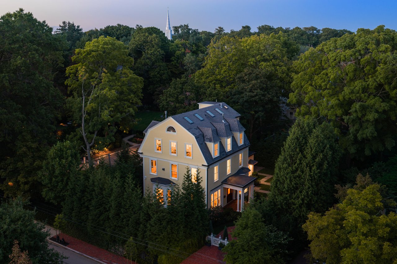 Aerial view of a yellow Victorian home with a mansard roof and glowing windows, surrounded by mature trees.