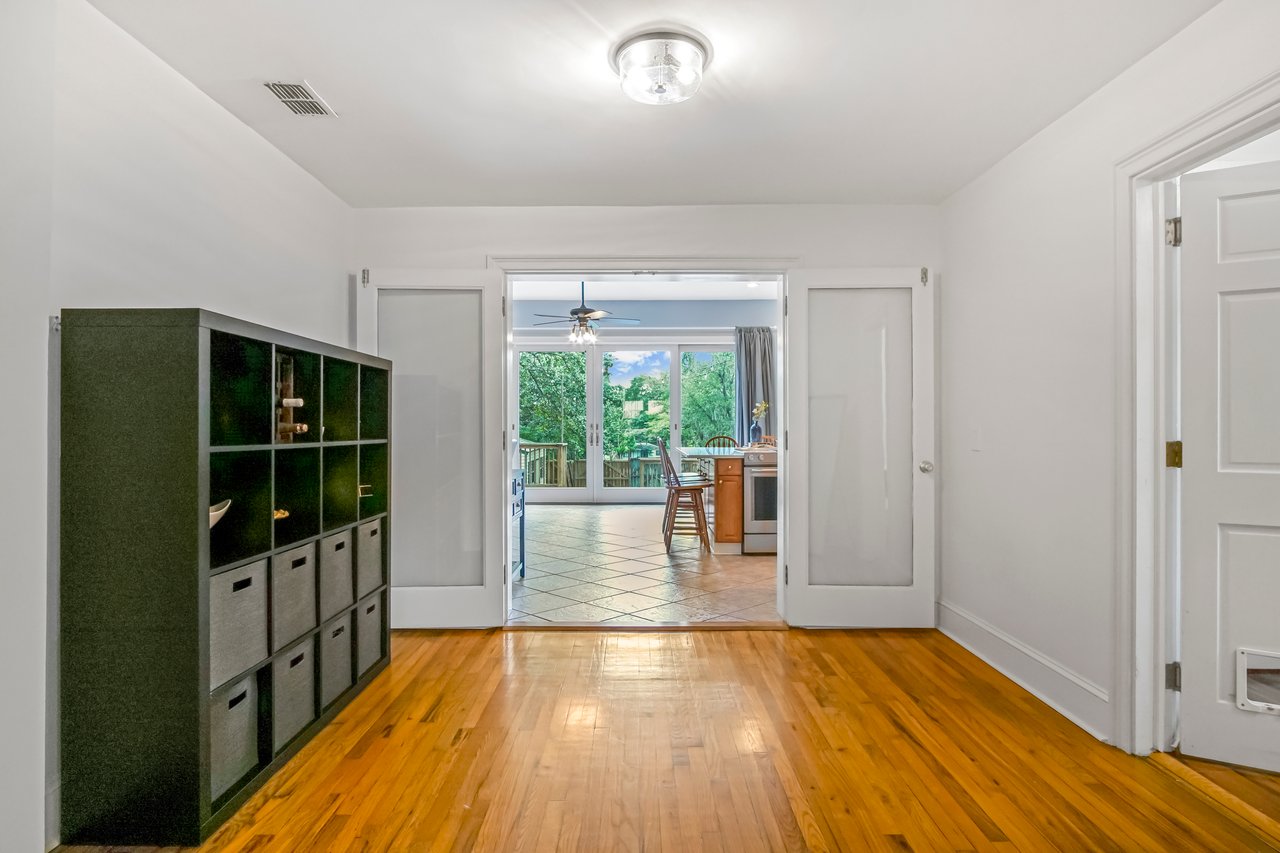 Bright hallway with wooden floors, white walls, and a storage unit on the left. Doors open to a room with a view of greenery, creating an inviting and airy feel.