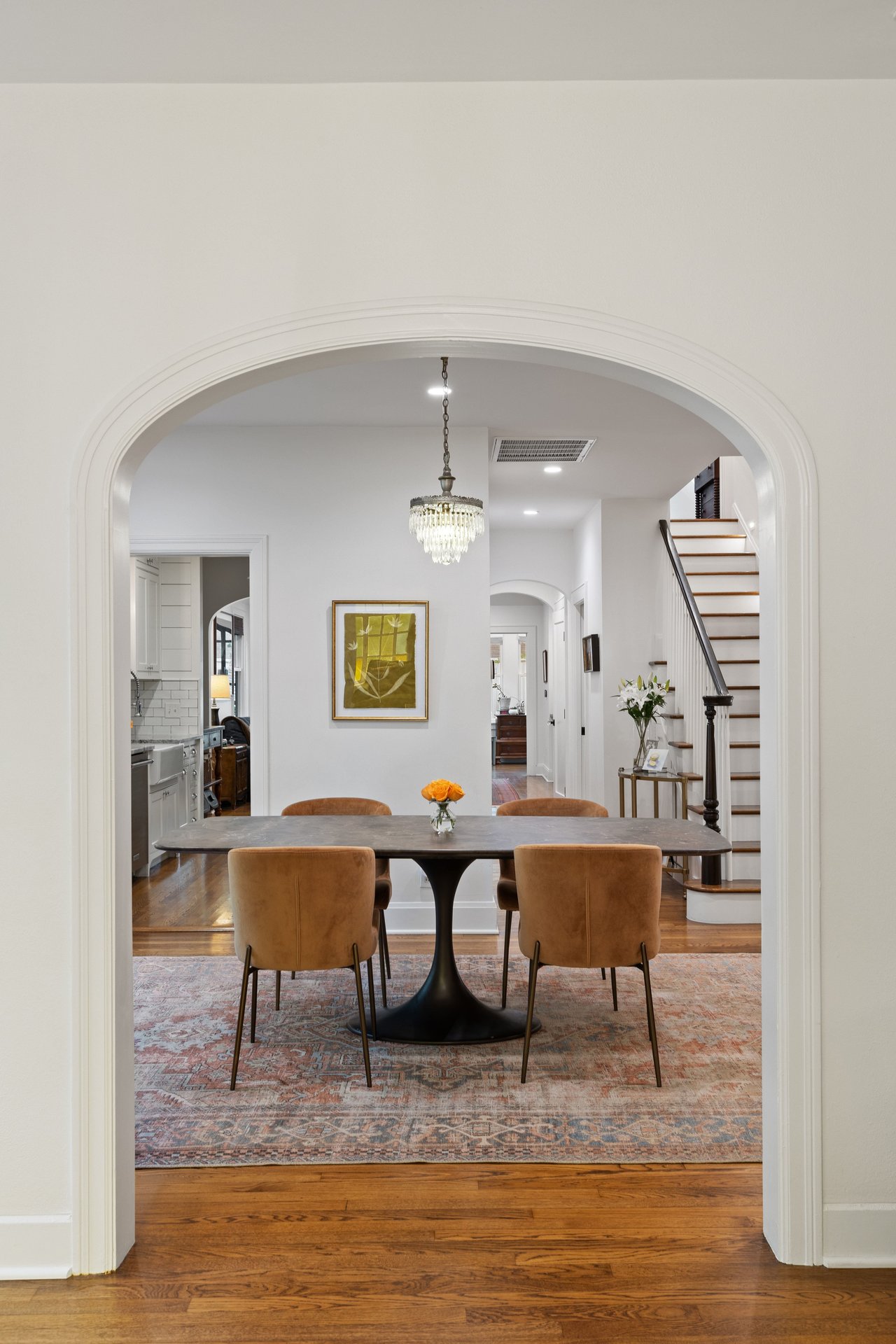 Dining room with a pedestal table, crystal chandelier, patterned rug, and staircase.