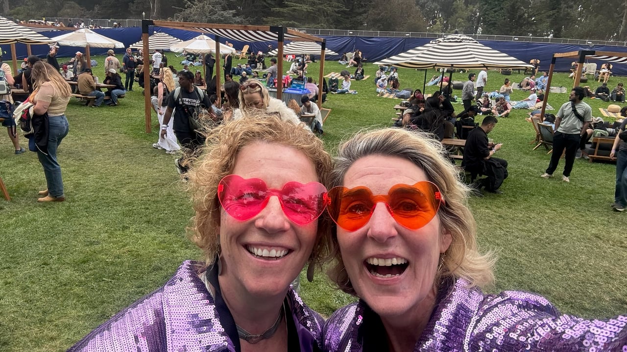 Two women smiling together at an outdoor daytime festival with picnic blankets and tents