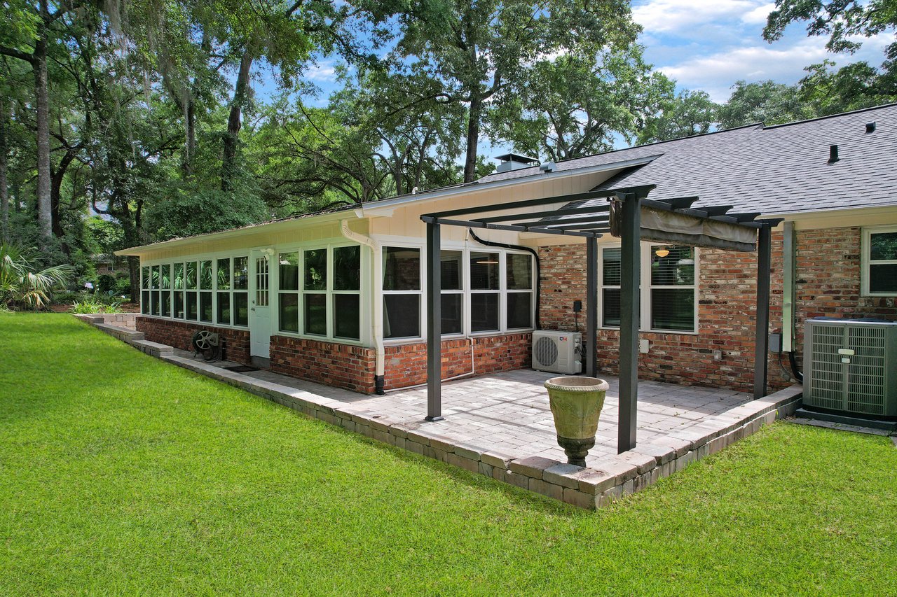 Rear view of a brick house with a sunroom featuring numerous windows. A pergola-covered patio with a decorative planter faces a lush green lawn.