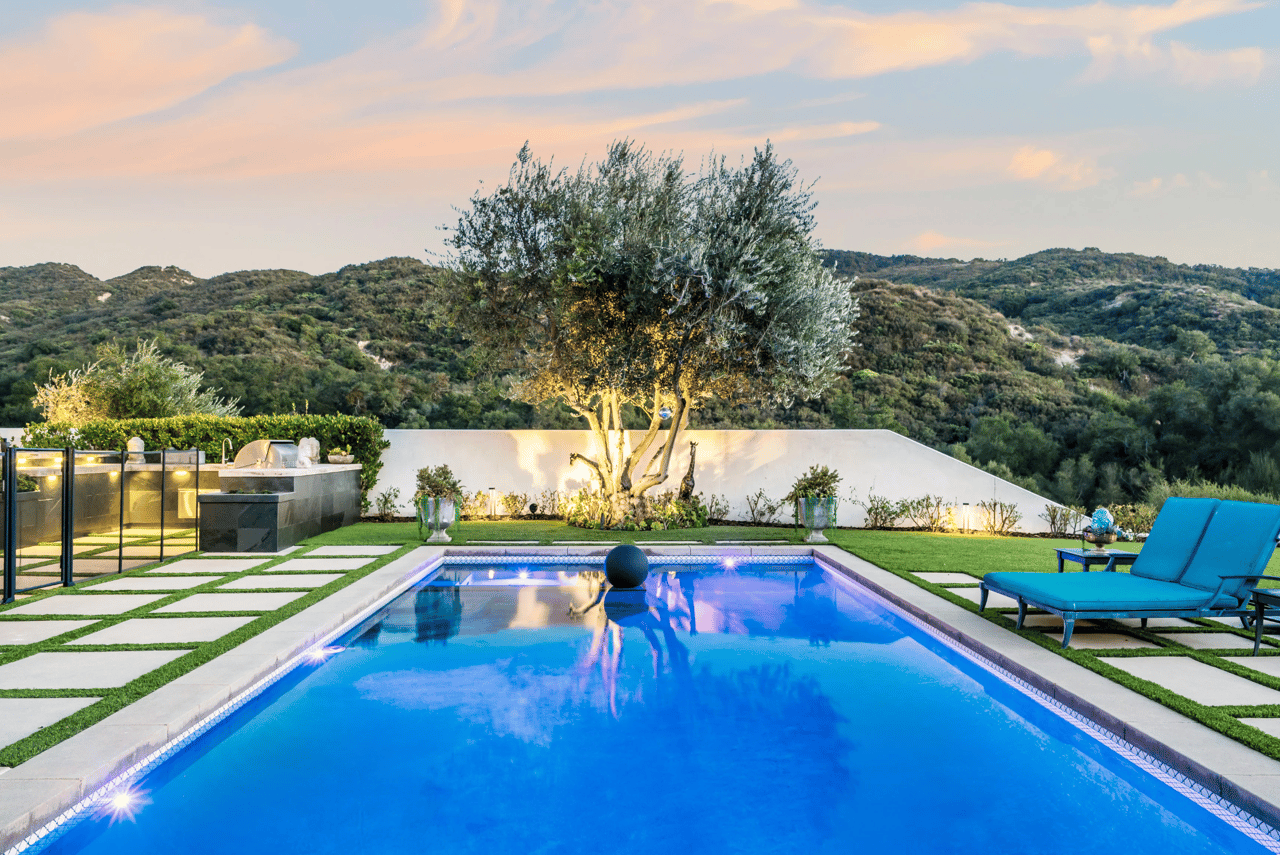 Backyard swimming pool with turquoise loungers, an outdoor kitchen, and wooded hills at sunset.