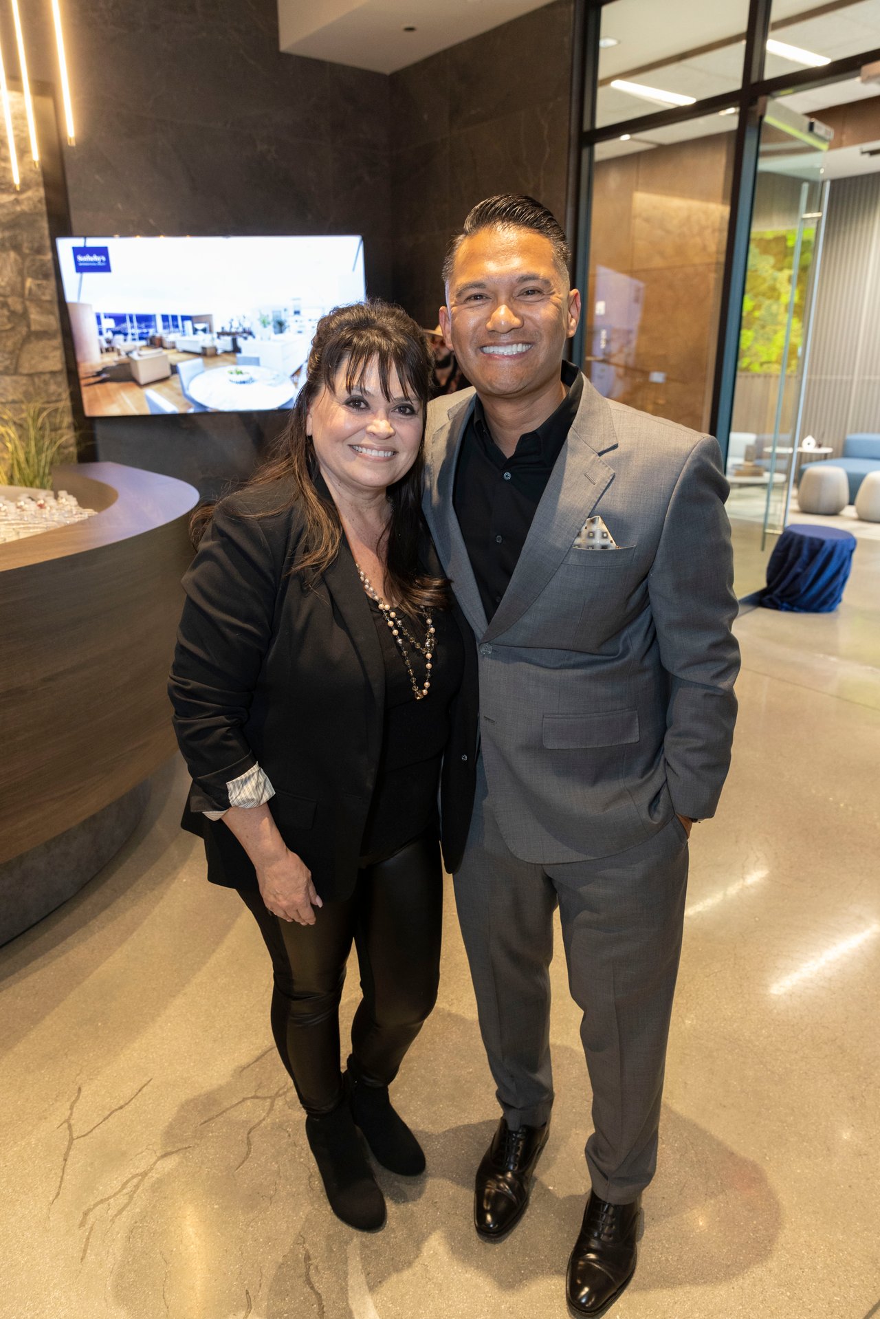 A woman in black and a man in a gray suit pose together inside a modern office.