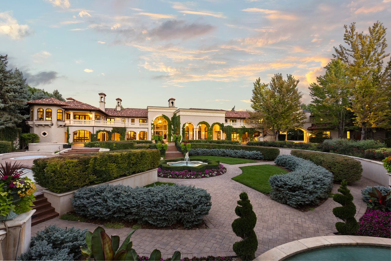 Estate gardens with clipped hedges, brick paths, a central fountain, and a Mediterranean-style home at sunset.