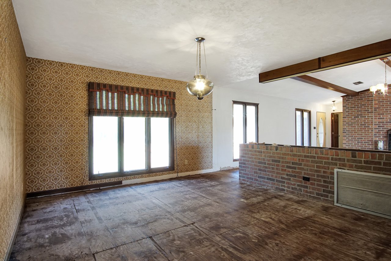 Spacious room with retro patterned wallpaper, large windows, and a brick half-wall. Wooden floors and ceiling beams create a warm, vintage atmosphere.