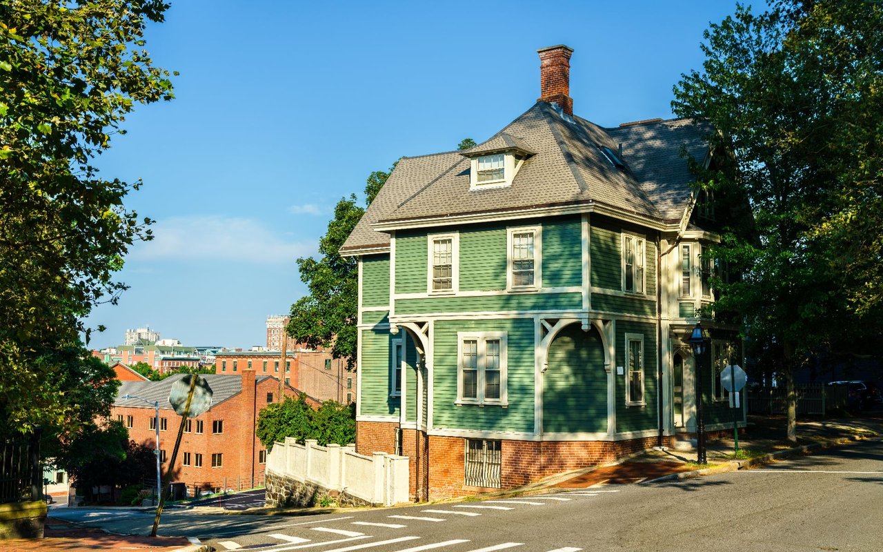 Green Victorian-style house with white trim, a brick foundation, and a tall chimney on a tree-lined street.