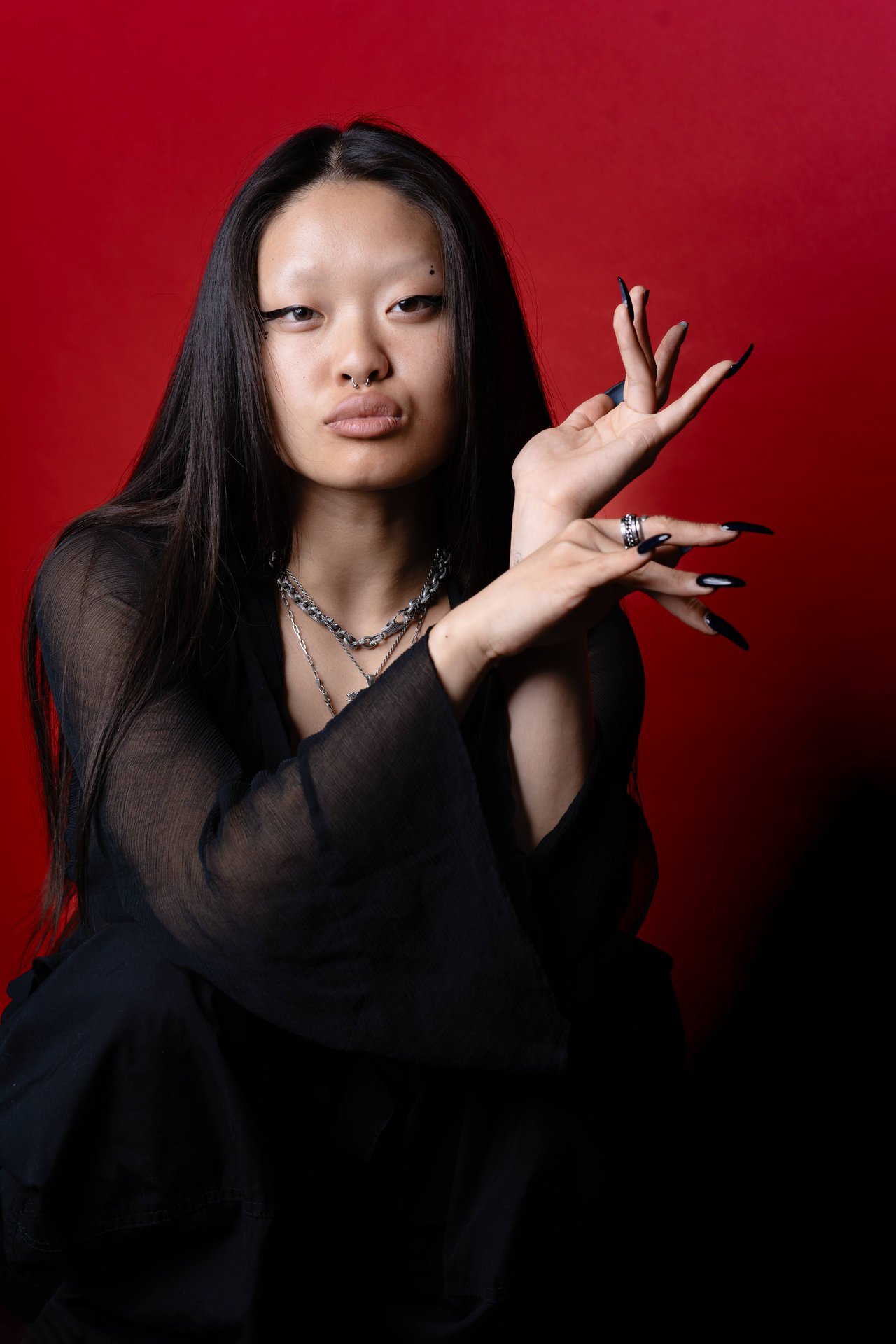 Dancer Jin poses against a red backdrop with raised hands, long pointed nails, and layered silver necklaces.