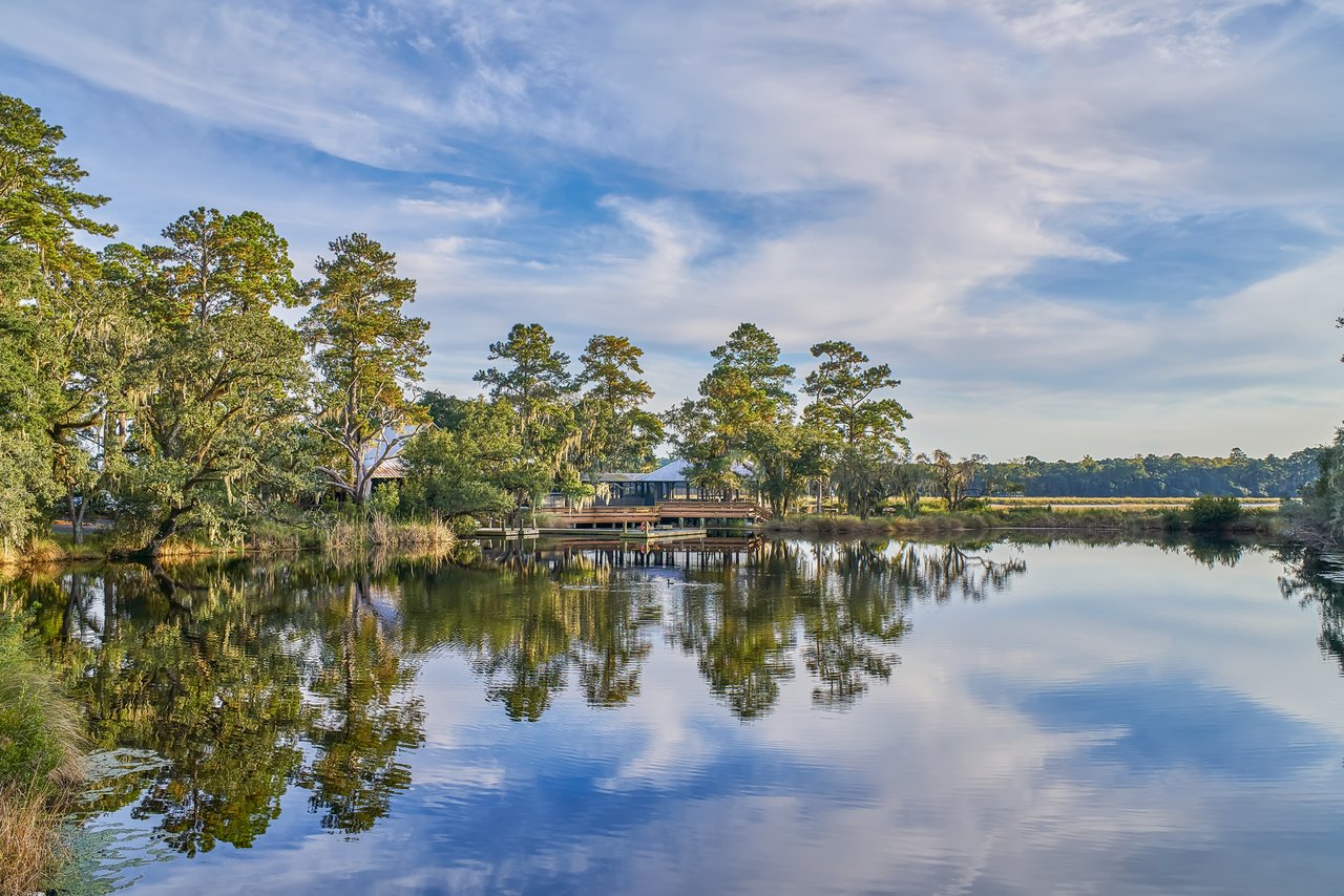 A calm pond reflects tall trees, a waterside building, and a dock beneath a partly cloudy sky.