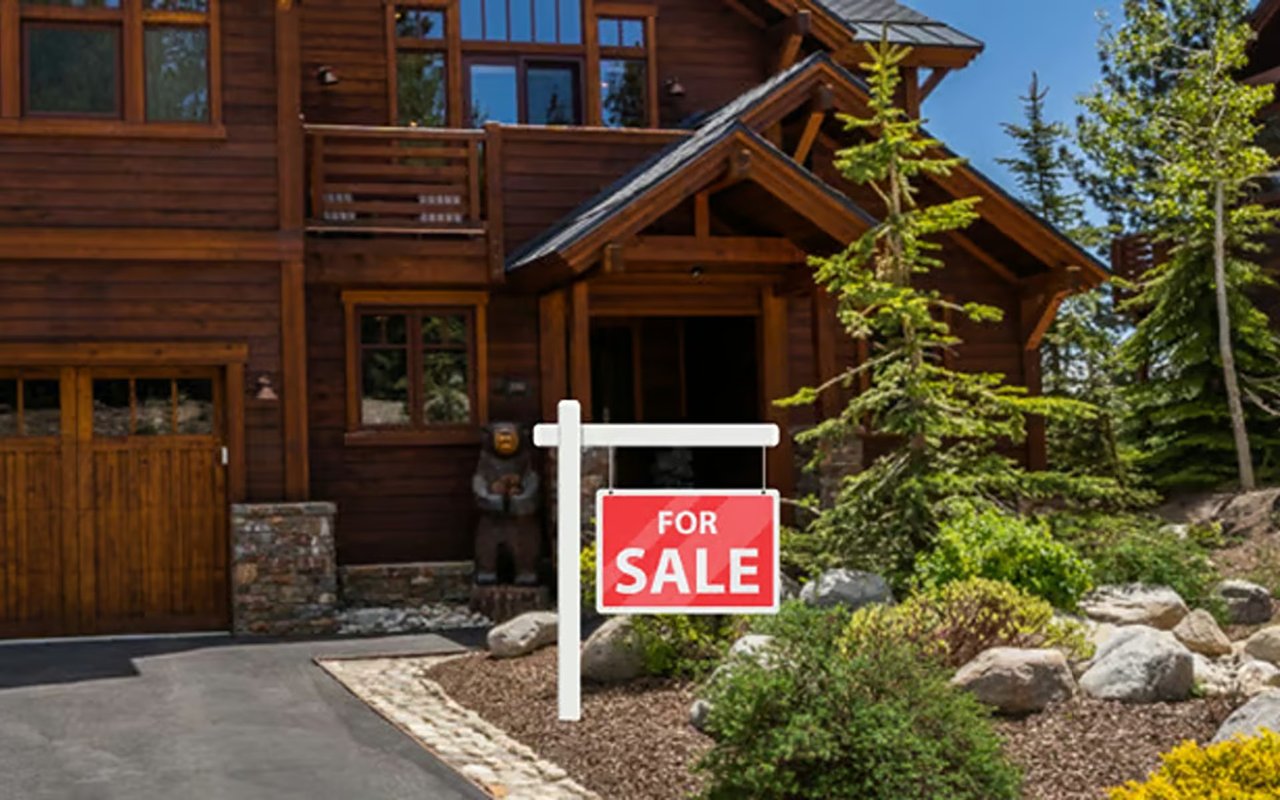 Dark-wood house among evergreens with a red FOR SALE sign beside the driveway.