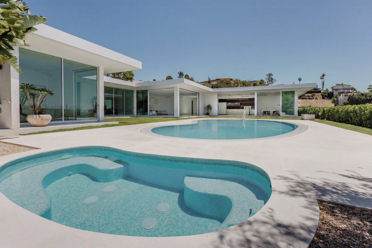 Modern glass-walled home behind two turquoise pools, including a curved pool with built-in seating.