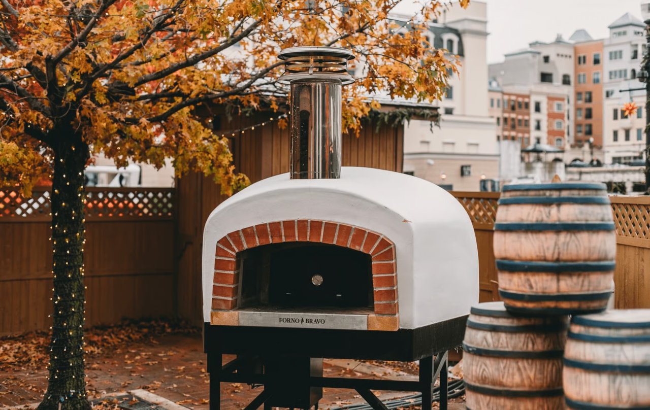 Luxury patio setup featuring a Forno Bravo masonry pizza oven, decorative whiskey barrels, a tree with string lights, and orange fall foliage with city buildings behind.