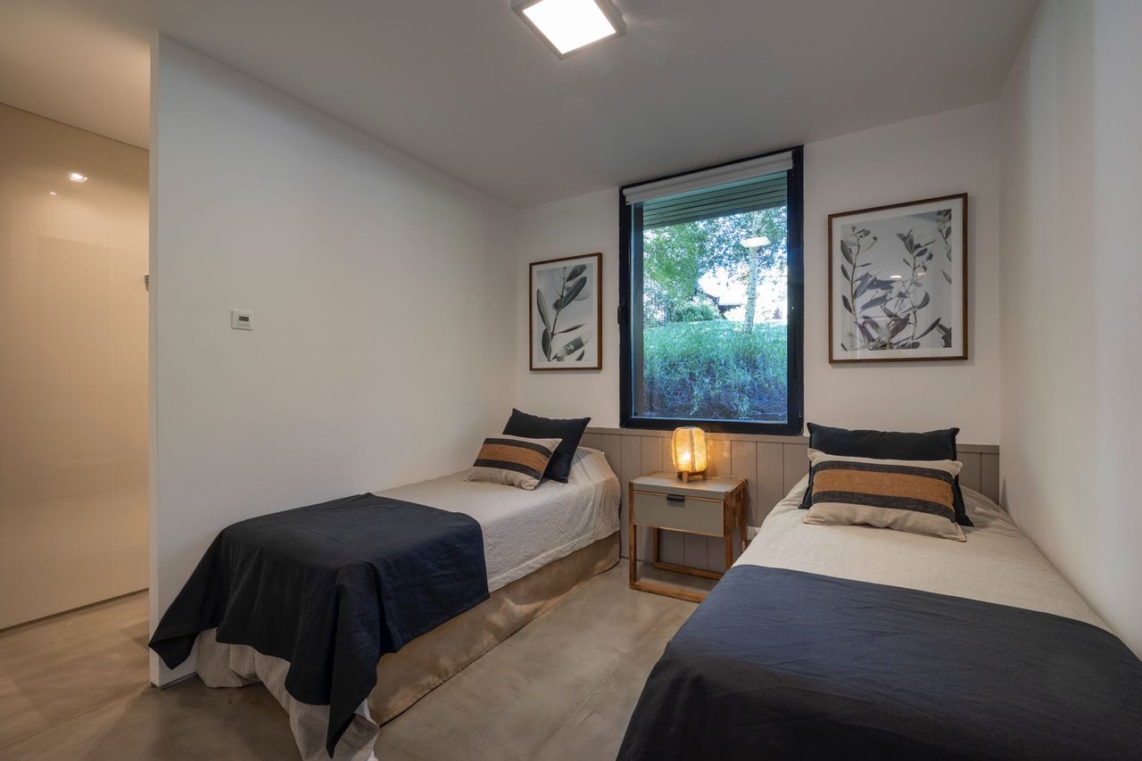 Twin bedroom with charcoal bedspreads, botanical prints, and a window overlooking greenery.