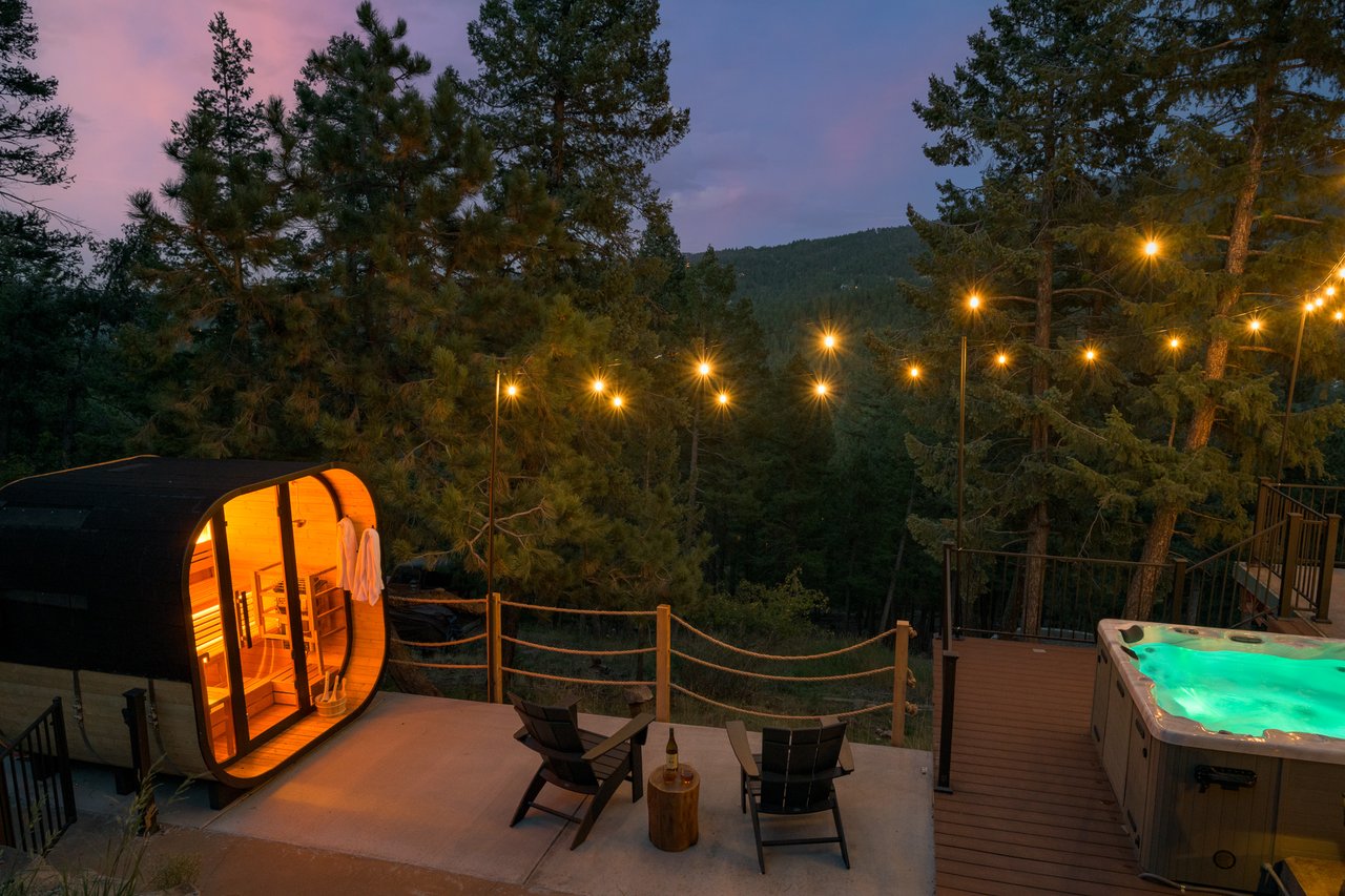 Woodland deck at dusk with a lit barrel sauna, hot tub, and string lights among the trees.