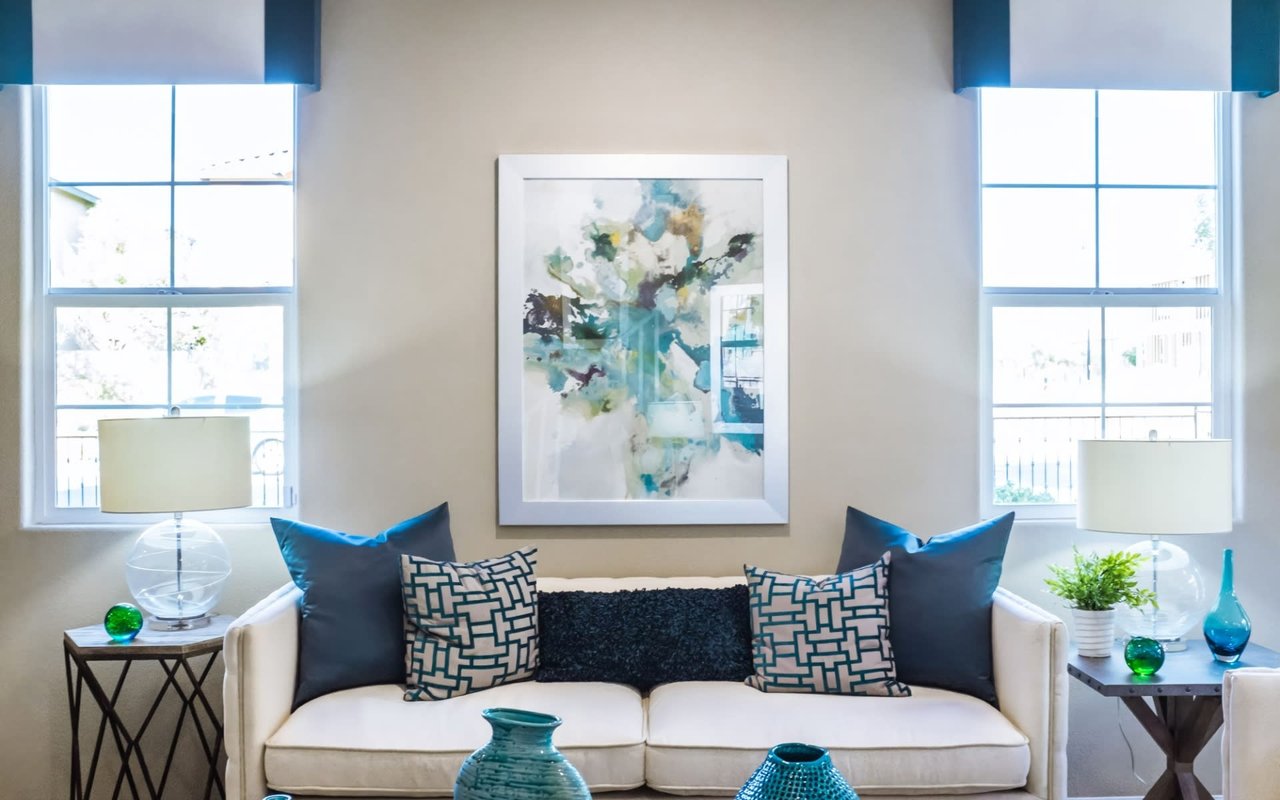 Staged living room showcasing a cream loveseat with satin teal pillows, a large silver-framed watercolor painting, geometric side tables, and glass orb table lamps.