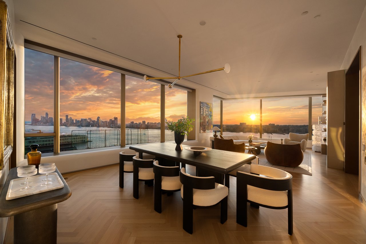 Furnished apartment dining area with floor-to-ceiling windows overlooking a river and city skyline at sunset.