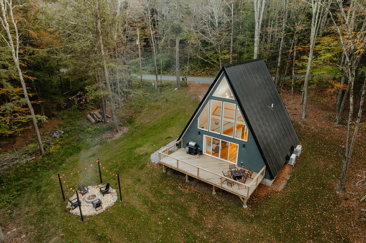 A-frame cabin with a broad deck and separate fire pit in a wooded autumn setting.