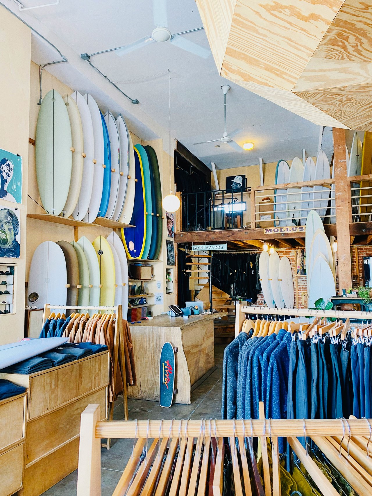 Surf shop interior with rows of surfboards, a wooden mezzanine, and clothing racks.