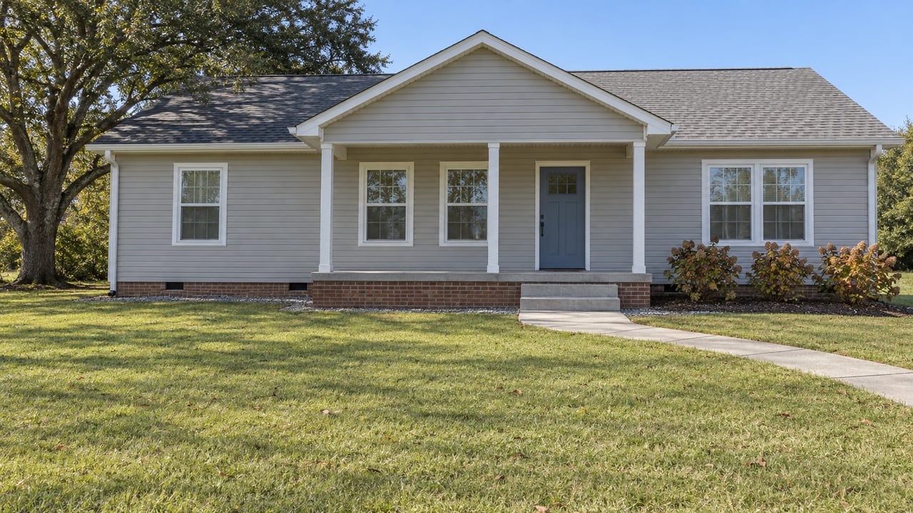A modest gray single-story house with a sheltered front porch, broad lawn, concrete walk and mature spreading tree.