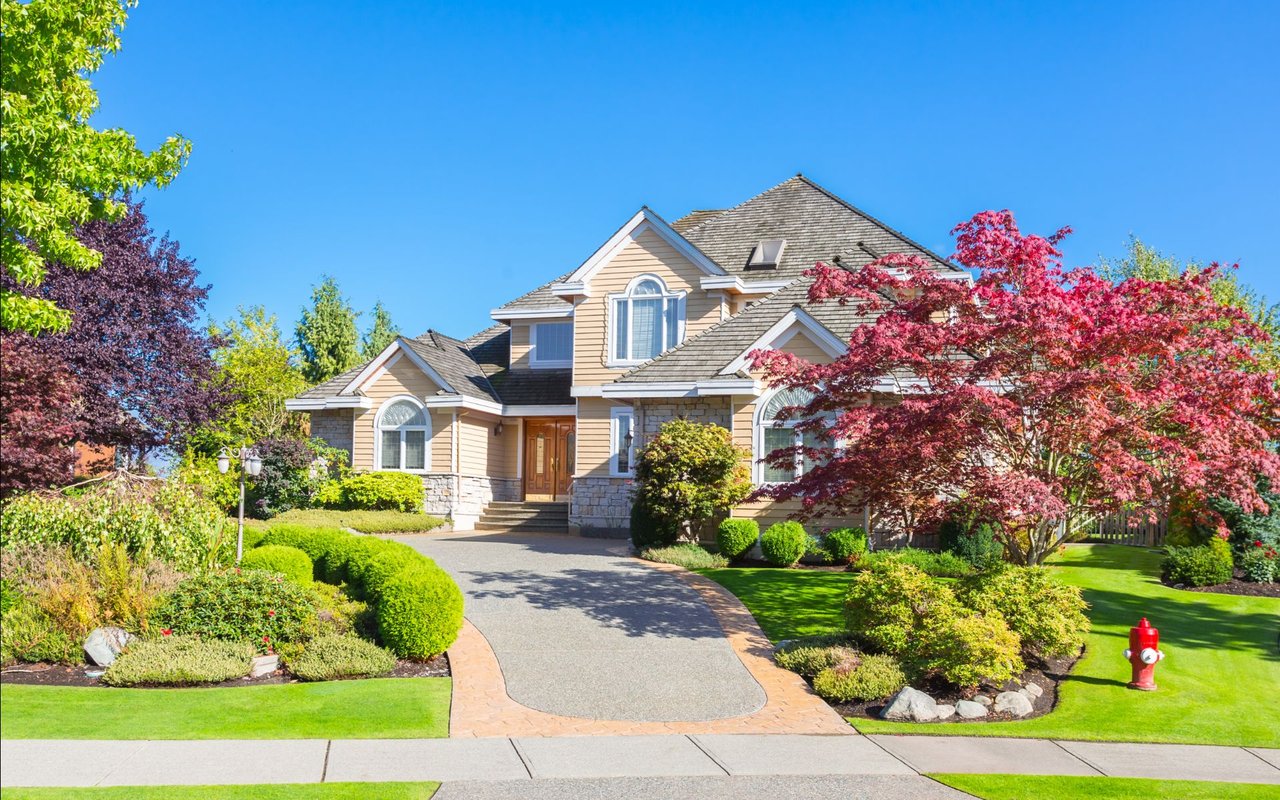 Two-story tan house with stone accents, a curved driveway, and mature colorful landscaping.