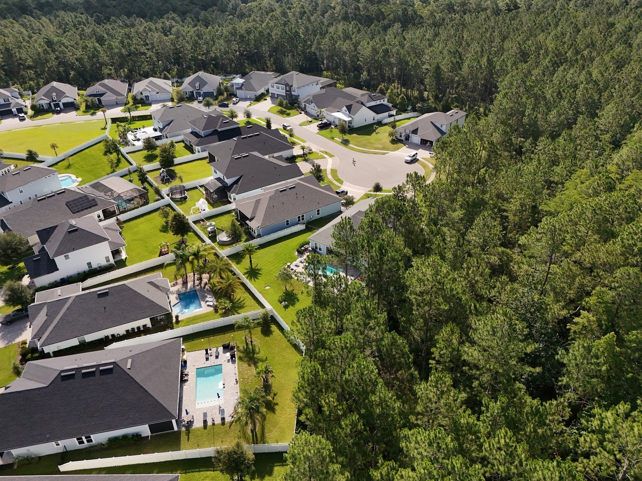 Aerial view of homes with fenced yards and pools beside dense woodland.